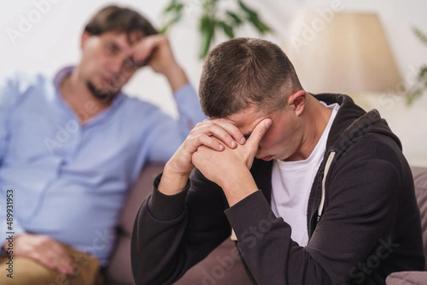 Fototapeta frustrated, young man is sitting at home on couch. difficult teenager covers his face with his hands, he has problems. father talks to his son, discusses problems, supports him. Transitional age
