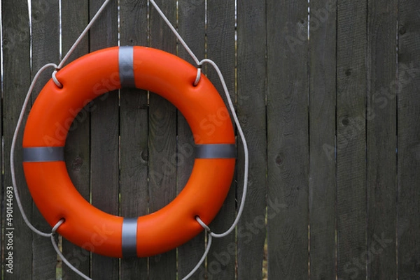 Fototapeta Orange lifebuoy hanging on grey wooden fence, space for text. Rescue equipment