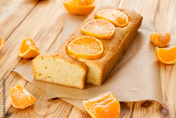 Obraz Moist orange fruit pound cake on parchment on rustic wooden background with slices of orange. Delicious breakfast, traditional English tea time. Reciepe of orange pie loaf.