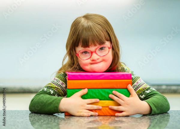 Fototapeta A child with down syndrome with glasses sitting at a table with books in an embrace