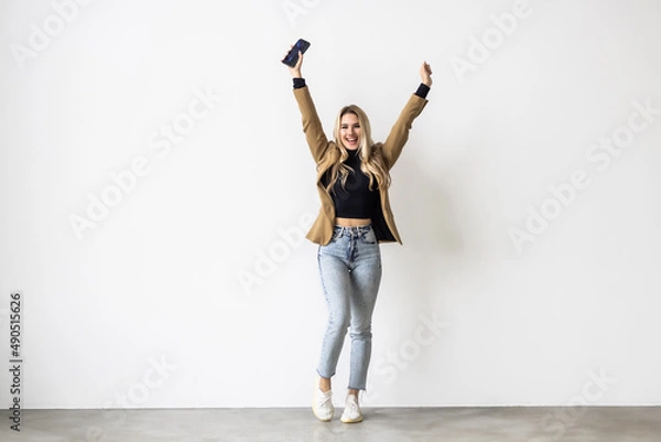 Fototapeta Satisfied smiling woman winning money prize on mobile phone, holding smartphone and fist pump with pleased face on white background