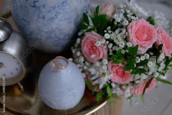 Fototapeta Wedding rings on a nightstand with a clock and a vase.
