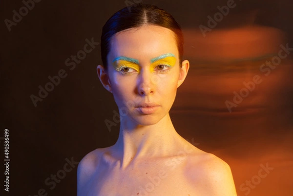 Fototapeta A woman's face in the blue and yellow national colors of Ukraine looks at the camera isolated on a dark background. Flag of UA, no war.
