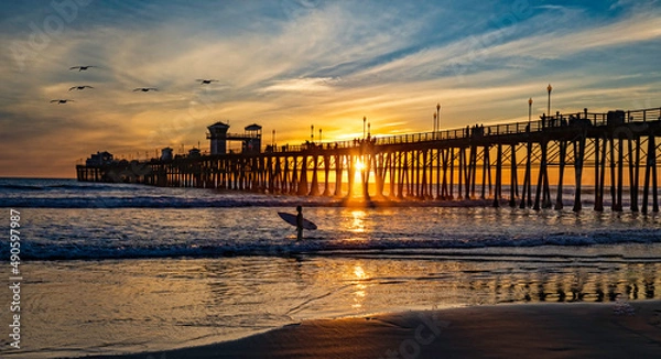 Obraz Surfer at Oceanside Pier. A colorful sunset image with reflections and pelicans passing.