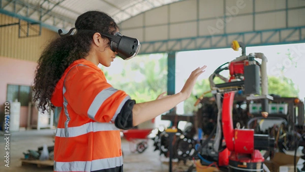 Obraz Young Africa American engineer is controlling Futuristic robotic arm with a virtual reality Headset. Testing robot by moving hand at factory. VR Technology industrial and modern equipment.