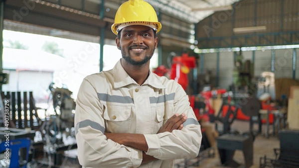 Obraz Close up portrait of confident Africa American engineer man wearing safety helmet standing in the robotic arm manufacturing. Looking forward and crossed arm. Technology and industrial concept..