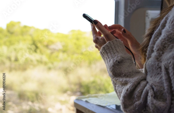 Fototapeta Horizontal image of a woman using her smart phone inside a camper van, with the door open to enjoy a beautiful day.