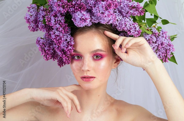 Fototapeta Portrait of a young girl in the studio with lilac flowers on her head, close-up. Creative beautiful make-up in doll style with lilac flowers on the head close-up.