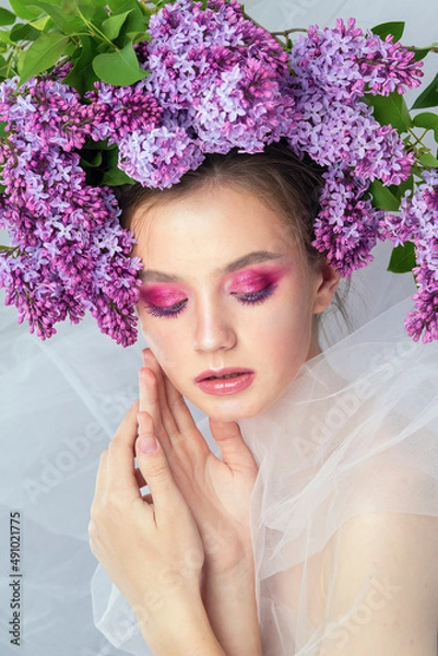 Fototapeta Portrait of a young girl in the studio with lilac flowers on her head, close-up. Creative beautiful make-up in doll style with lilac flowers on the head close-up.