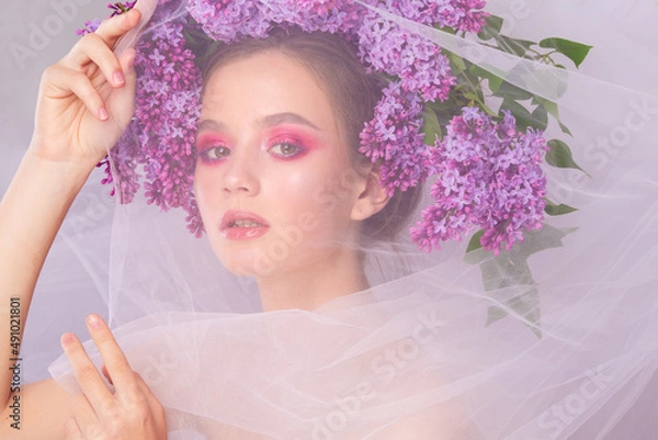 Fototapeta Portrait of a young girl in the studio with lilac flowers on her head, close-up. Creative beautiful make-up in doll style with lilac flowers on the head close-up.