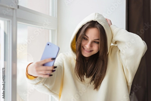 Fototapeta Happy female blogger taking selfie portrait on smartphone camera for social media posting, cheerful teenage girl talking on video call with family or friends via mobile phone.Soft selective focus.