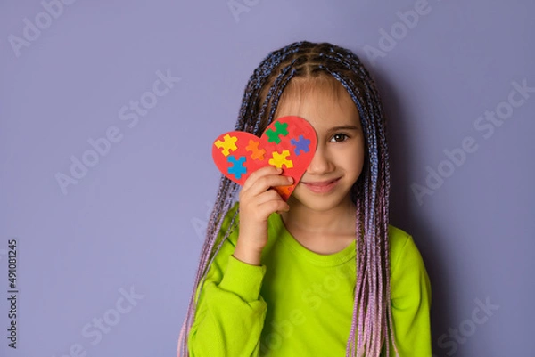 Obraz A girl with blue-purple Afro pigtails in a light green top holds a heart with puzzle pieces in her hands as a sign of solidarity with people suffering from autism spectrum disorder. World Autism day