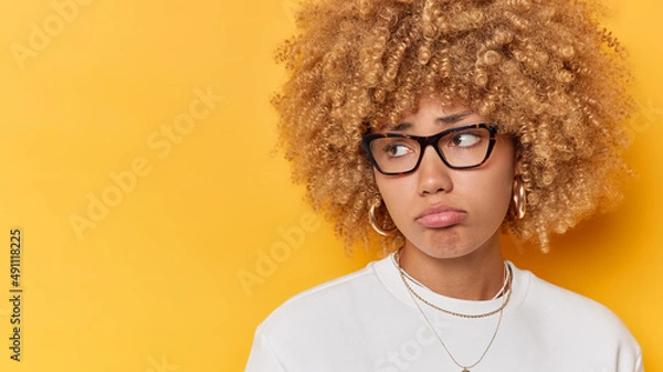 Obraz Horizontal shot of discontent woman with curly hair purses lips looks sadly away expresses negative emotions has troubles isolated over yellow background blank space for your promotional content