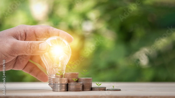 Fototapeta Man holding light bulb and many growth stack coins with plant in front. concept saving money for finance accounting and idea for growing saving money.