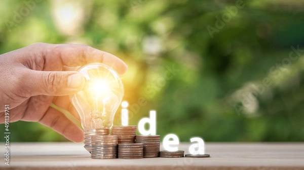 Fototapeta Man holding light bulb and many growth stack coins with plant in front and with the word Idea. concept saving money for finance accounting and idea for growing saving money.