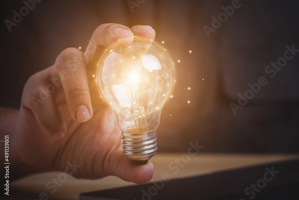 Fototapeta Businessman hand holding light bulb and using laptop on wooden table. new idea creativity concept with innovation and inspiration.