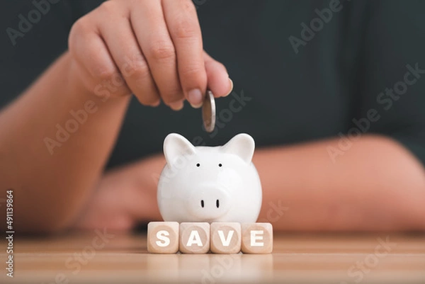 Fototapeta Close-up woman's hand holding a gold coin in a white piggy bank on wood block with Word save. Save money and financial investment.