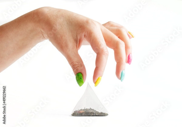 Fototapeta Triangular tea bag-pyramid and woman fingers with multi-colored manicure on a white background. Green tea with fruits and berries, flavoring herbs. Selective focus