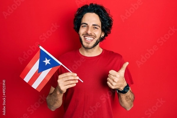 Obraz Handsome hispanic man holding puerto rico flag smiling happy and positive, thumb up doing excellent and approval sign