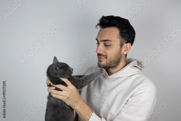 Fototapeta Young man holding cat on gray isolated background. Care and maintenance of pets.	