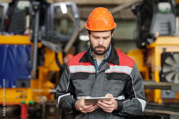 Fototapeta Serious young foreman or technician in safety helmet and uniform scrolling through online technical information in tablet
