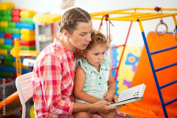 Fototapeta Teacher reads a book with a magical story to a little girl in kindergarten
