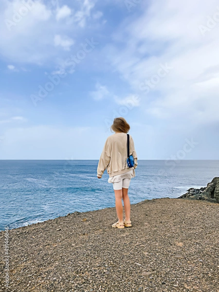 Fototapeta One young woman seen from back wearing white and beige clothes standing on a rock cliff on a windy day on blue sky and sea background. Travelling concept. Well Being and peacefulness.