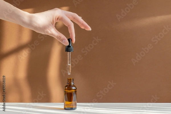 Obraz Pipette with cosmetic serum in women's hands on light brown background, in rays of sunlight. Concept of body care, rejuvenation. Close-up, copy space