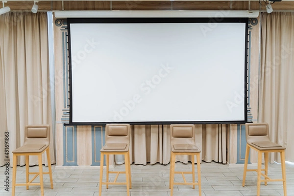 Fototapeta A white screen for showing the video hangs on the wall. Four chairs stand on the stage. Conference hall, business class for performances.