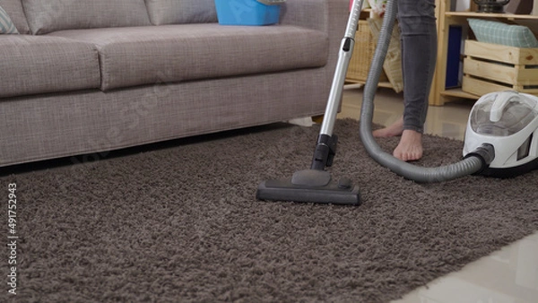 Obraz close up view vacuum cleaner head moving back and forth on the gray rug by the sofa with a woman standing barefoot in the background.