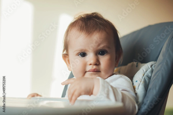 Fototapeta Caucasian baby girl sitting in high chair and looking curious into the camera. Baby care. Morning meal.