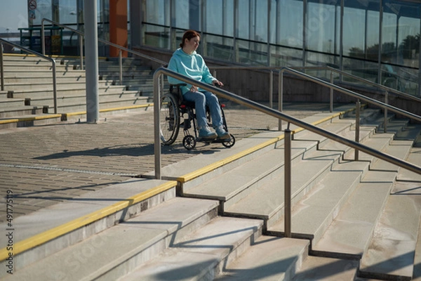 Fototapeta A caucasian woman in a wheelchair cannot go down stairs without a ramp. Hard to access environment. 