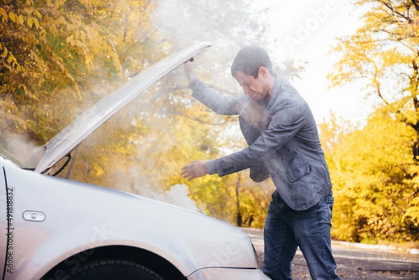 Obraz A man looks under the open hood of a car. The car broke down on the road. The engine is smoking
