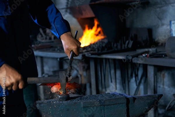 Fototapeta Close up worker in blue uniform keeping hammer and creating nice decorations for fence in large modern smithy. Concept of process creating decorations in professional workshop.