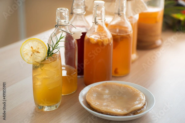 Obraz Kombu tea in a glass with ice, lemon slices and rosemary leaves, Homemade fermented raw kombucha tea in bottles and glass jars mix with fruit juice and scoby on table.Healthy natural probiotic drink.