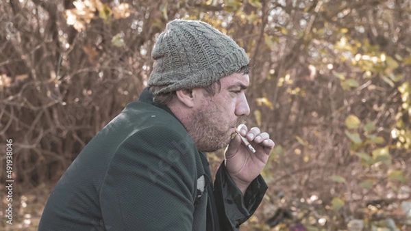 Obraz a poor homeless man with dirty hands smokes a cigarette in modern society. Economic recession, unemployment, poverty, hunger, pension, global crisis, the concept of the problem of inequality