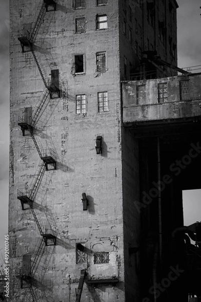Fototapeta Black and white street photo with the charm of the ugly architecture of old factory buildings. The concrete structure of the plant with a metal staircase and different windows. Conceptual photo with a