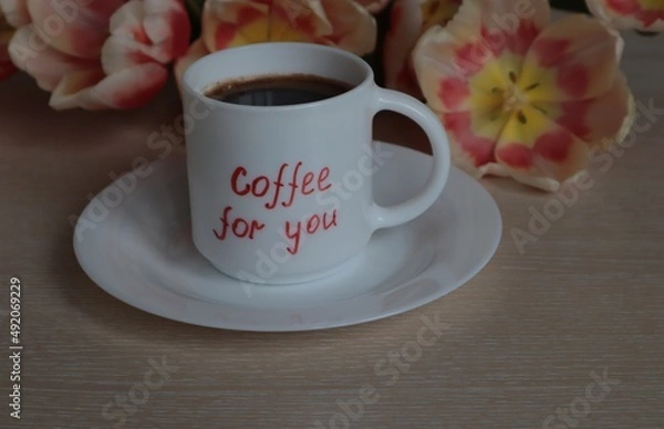 Obraz A white mug with black coffee stands against a background of tulips on a light surface.
