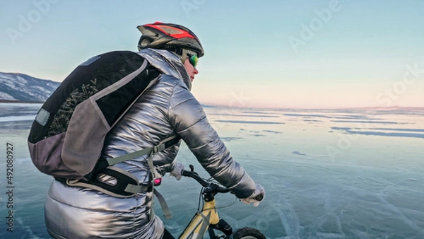 Fototapeta Woman is riding bicycle on the ice. Girl is dressed in a silvery down jacket, cycling backpack and helmet. Ice of the frozen Lake Baikal. Tires on bike are covered with spikes. Traveler is ride cycle.