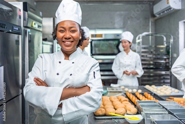 Fototapeta Smiling african  female bakers looking at camera..Chefs  baker in a chef dress and hat, cooking together in kitchen.Team of professional cooks in uniform preparing meals for a restaurant in  kitchen.