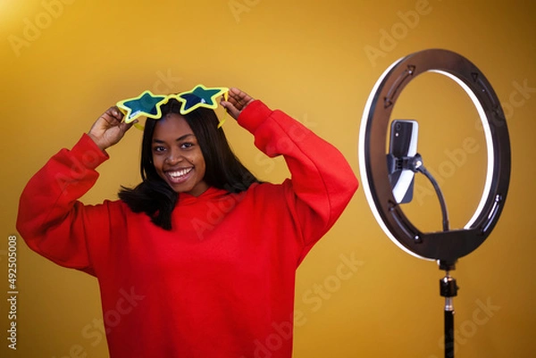 Obraz blogging, technology, emotions and people concept - a happy African-American vlogger girl in a red hoodie and big star glasses with a ring lamp and phone looking at the camera on a yellow background