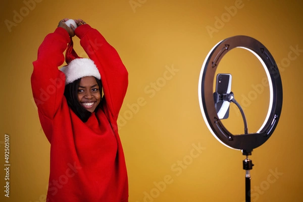 Obraz laughing african-american girl blogger in a red hoodie and a Santa hat with a lighted ring lamp and phone holding a pompom on a yellow background with copy space