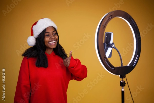 Obraz smiling african-american girl vlogger in a red hoodie and a Santa hat with a lighted ring lamp and phone showing thumbs up on a yellow background with copy space
