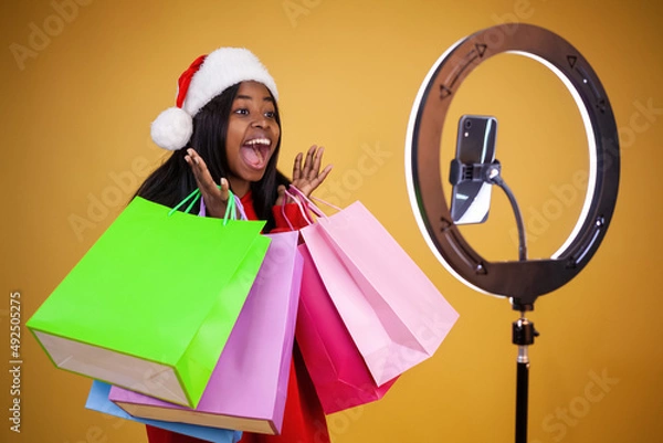 Obraz an admiring African-American girl in a red hoodie and Santa hat with large colorful bags, a ring lamp, and a phone on a yellow background with copy space