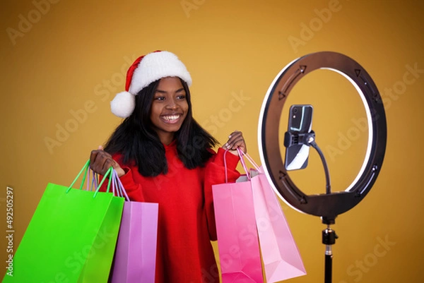 Obraz blogging, new year, technology, shopping and people concept - smiling African-American girl in a red hoodie and Santa hat with large colorful bags, a ring lamp and a phone on a yellow background