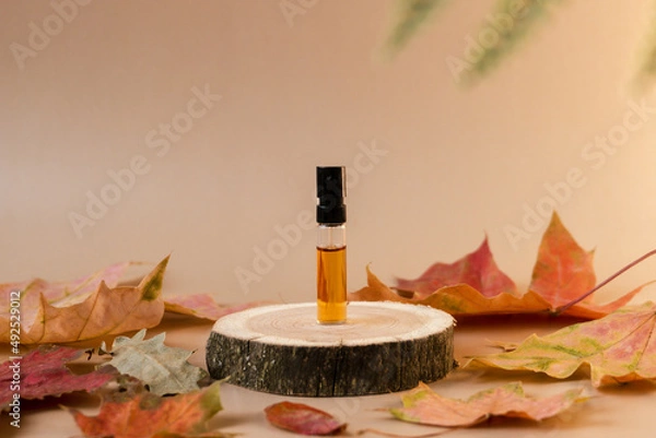 Fototapeta Glass perfume sample with brown liquid on a wooden tray standing on beige background with fallen leaves around. Luxury and natural autumn cosmetics presentation. Tester on a woodcut. Front view