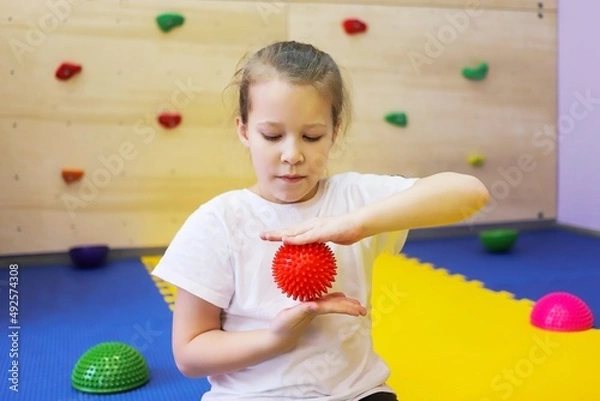 Fototapeta a little girl in white holds a red ball with spikes and massages her hands in a children's center so as not to get sick