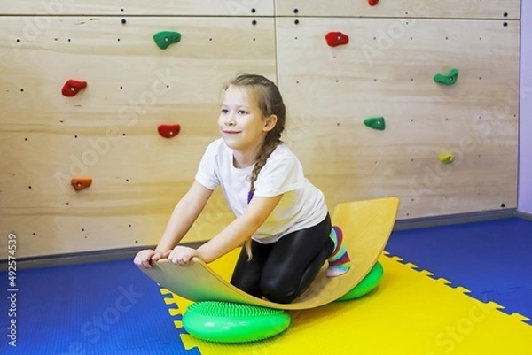 Fototapeta a little girl in white swings on a special balance beam to the sides in a children's center so as not to get sick