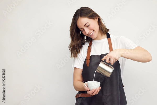Fototapeta Professional smiling barista woman in studio serving coffee, pour milk from a jug in a coffee cup. Café latte. Isolated on white background. 