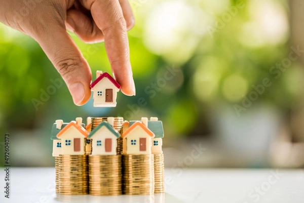 Obraz Woman hand hold for select a home model on the stack gold coin on sunlight in the public park, Loan for real estate or saving money for buy a new house to family in the future concept.